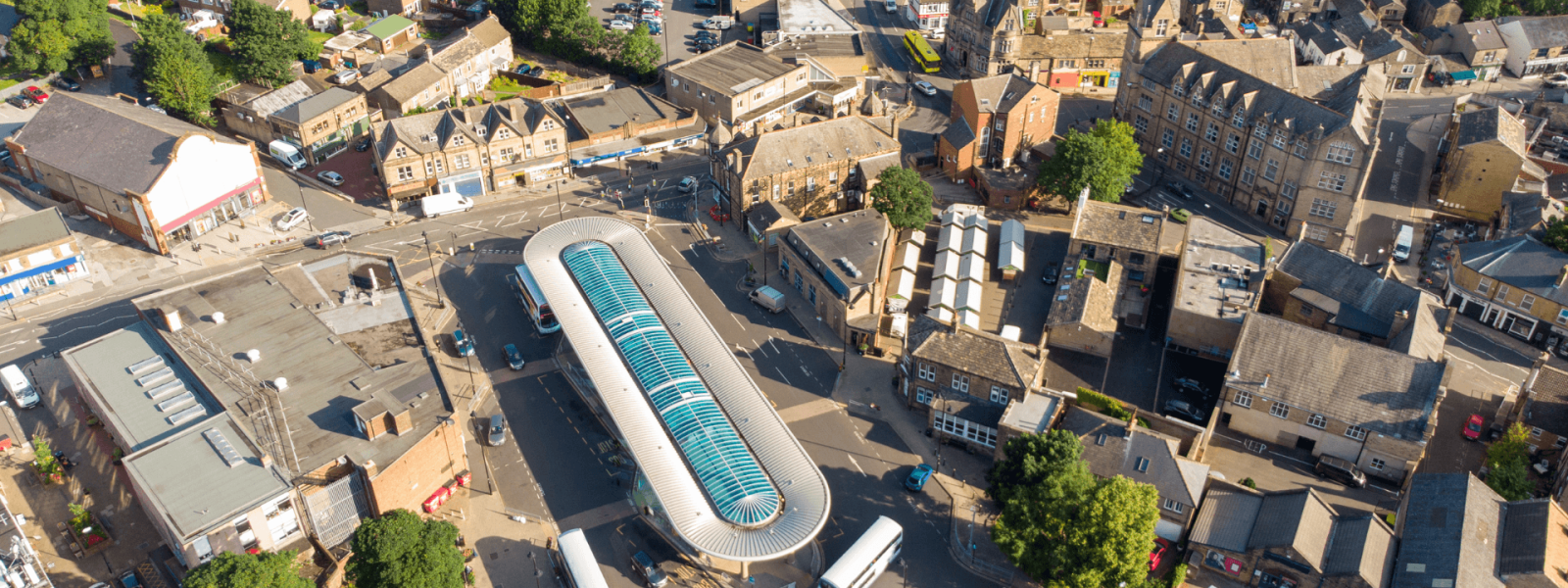 Aerial view of the town center of Pudsey, West Yorkshire on a sunny day. The image shows a mix of old stone buildings and newer structures. A distinctive modern curved glass roof building is visible in the center, likely a public building or transportation hub. The streets form a small roundabout in the middle of the scene. Parked cars line the streets and small green spaces with trees break up the urban landscape. The density and variety of buildings suggests this is the commercial heart of the town.