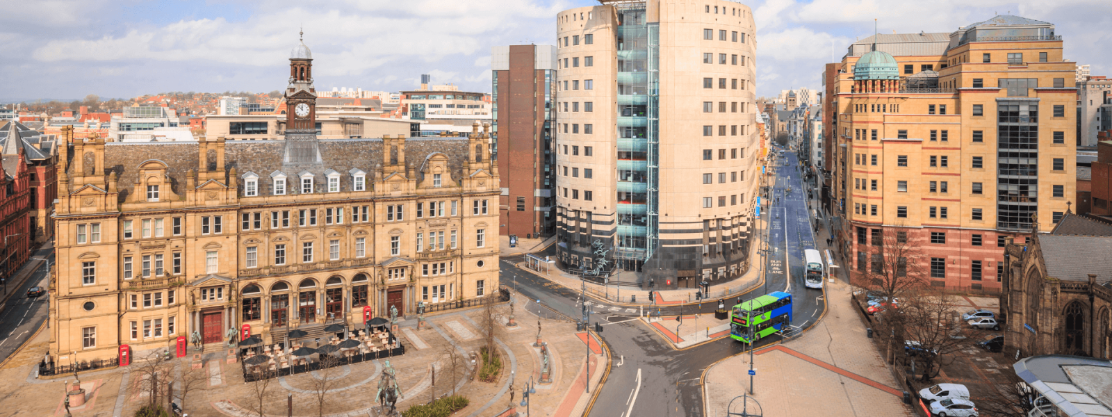 Aerial view of a city square in Leeds, England on a sunny day. The square is surrounded by historic stone and brick buildings, including a ornate clock tower. Modern glass and concrete high-rise buildings are also visible in the background. The square itself features a roundabout with streets radiating out, green spaces with trees, pedestrian areas, and a couple of green double-decker buses.