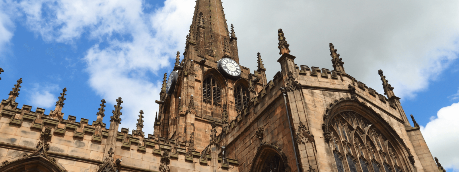 A large, ornate cathedral with a tall spire and numerous decorative spires and arches around the exterior. The cathedral appears to be constructed from sandstone, giving it a warm, golden hue against the partly cloudy blue sky. The architectural style suggests it is a Gothic cathedral, with intricate stone carvings and details adorning the facade.